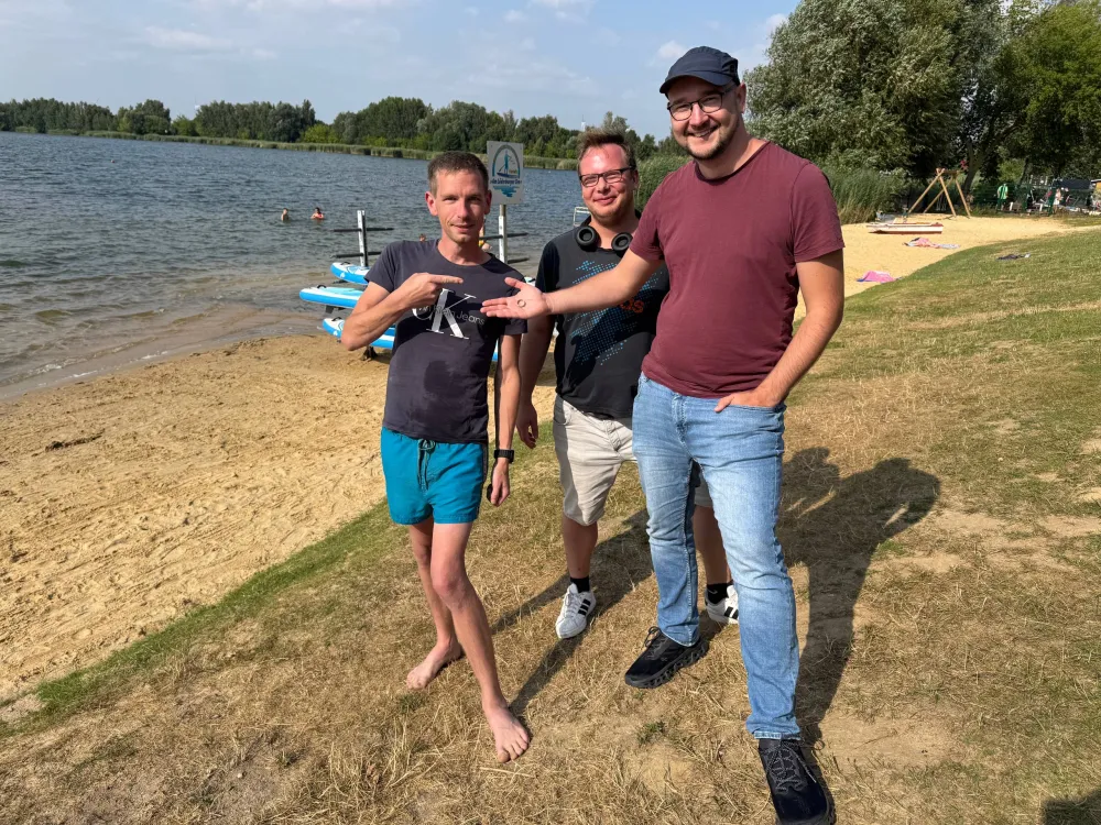 Drei Männer am sandigen Ufer des Löderburger Sees, einer zeigt auf einen kleinen Ring auf seiner Handfläche.