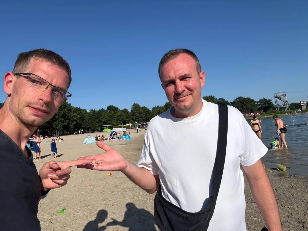 Zwei Männer am Sandufer des Tankumsees, einer zeigt auf zwei kleine Ringe in der geöffneten Handfläche des anderen.
